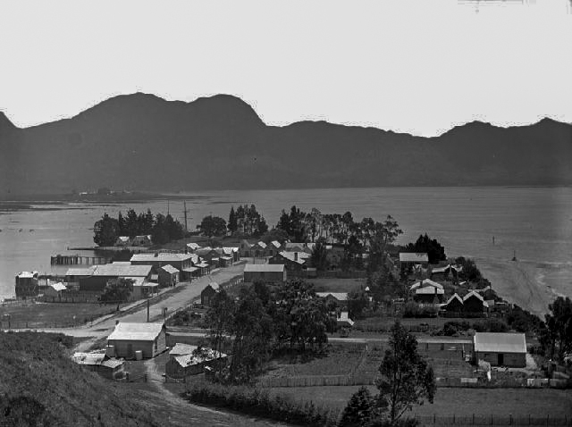 A historic black-and-white view of Collingwood, with clustered cottages and buildings along the waterfront beneath a backdrop of rugged hills.