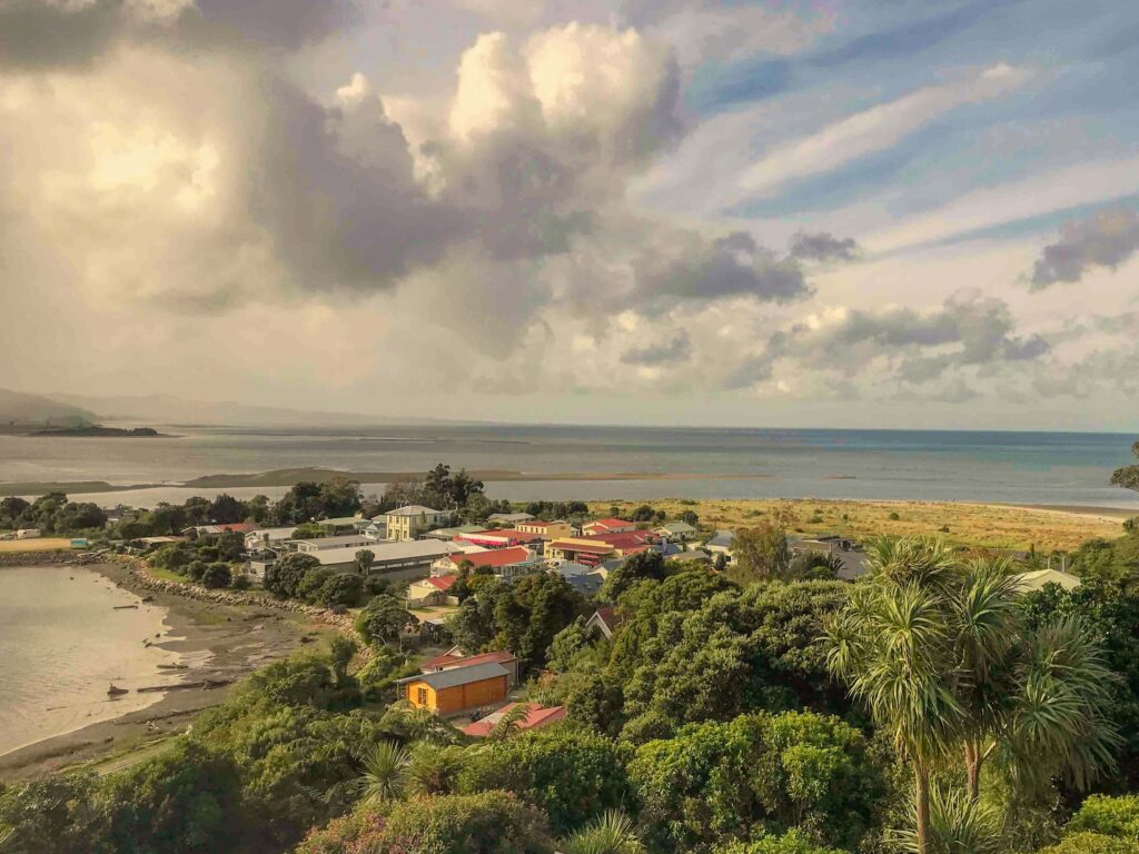 A seaside settlement with red, yellow, and white rooftops surrounded by lush greenery, overlooking the ocean with clouds and sunlight breaking through the sky.