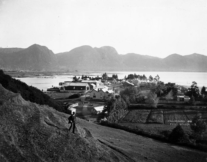 A historic photograph of Collingwood in 1921, showing a small coastal town framed by farmland, calm waters, and the rugged silhouette of surrounding hills.