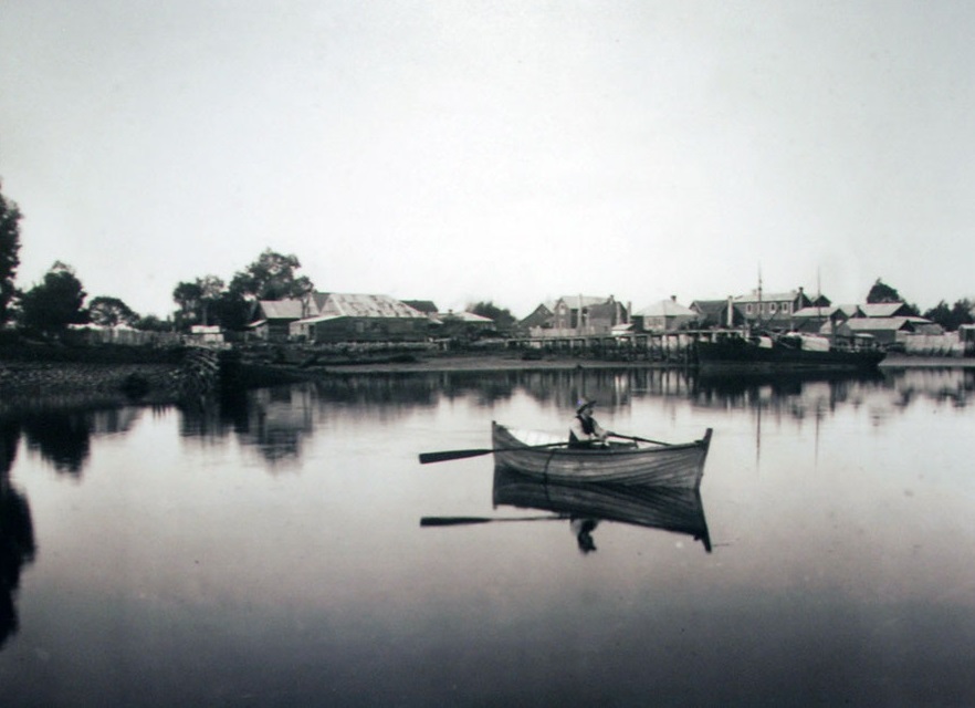 A lone rower glides across calm waters with the early township of Collingwood visible along the shore