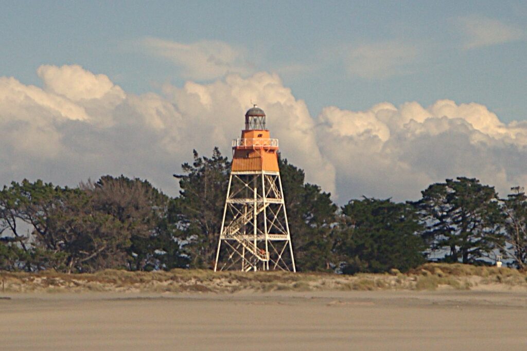 A tall metal-framed lighthouse with an orange top, set against trees and a cloudy sky at Farewell Spit.