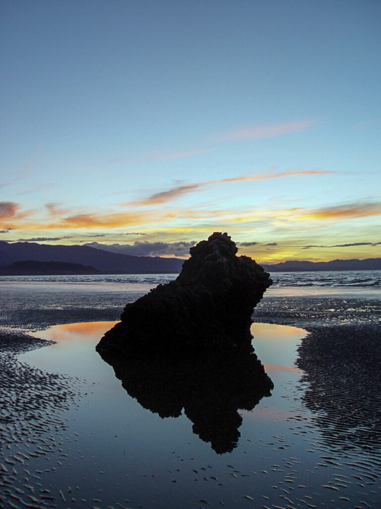 A lone rock stands reflected in a tidal pool, silhouetted against a pastel sunset sky.