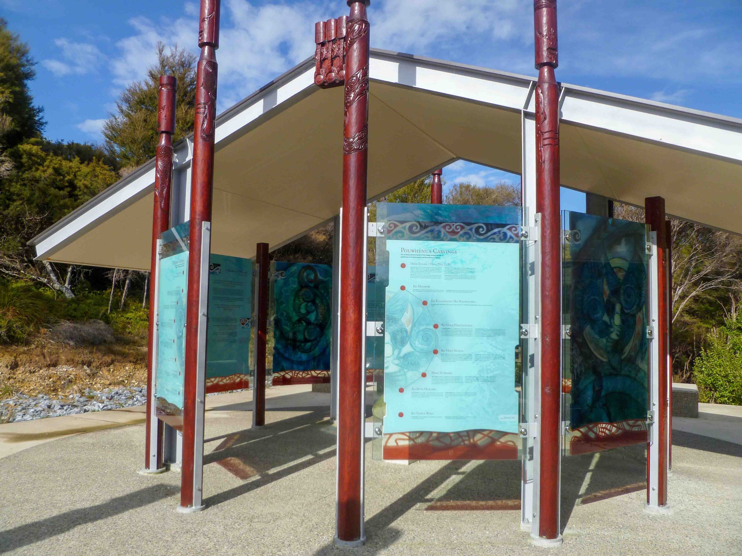 Entrance pavilion at Te Waikoropupū Springs, featuring carved red wooden posts and glass information panels with Māori cultural designs.