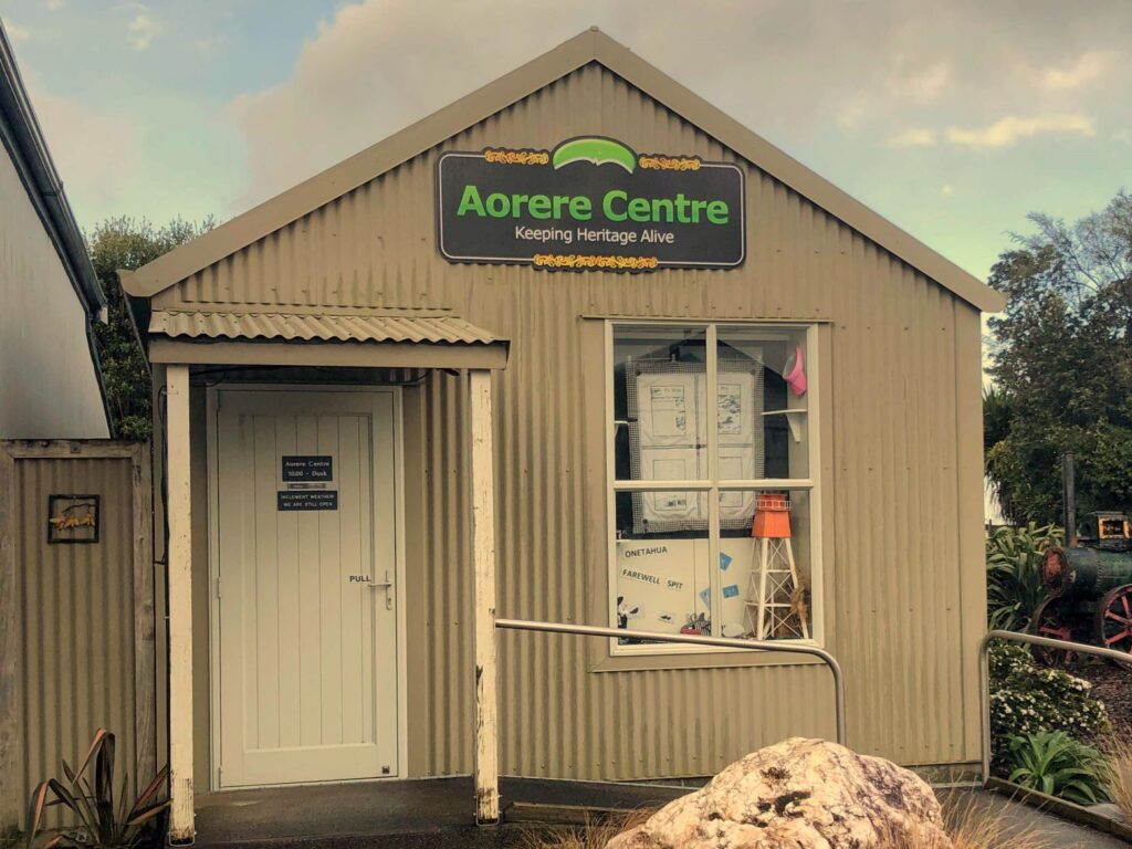 Front view of the Aorere Centre in Golden Bay, New Zealand, featuring a beige corrugated building with a sign reading “Aorere Centre, Keeping Heritage Alive.”