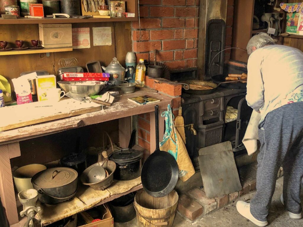 Heritage kitchen scene with a woman cooking on a cast-iron wood stove surrounded by vintage cookware and baking supplies.
