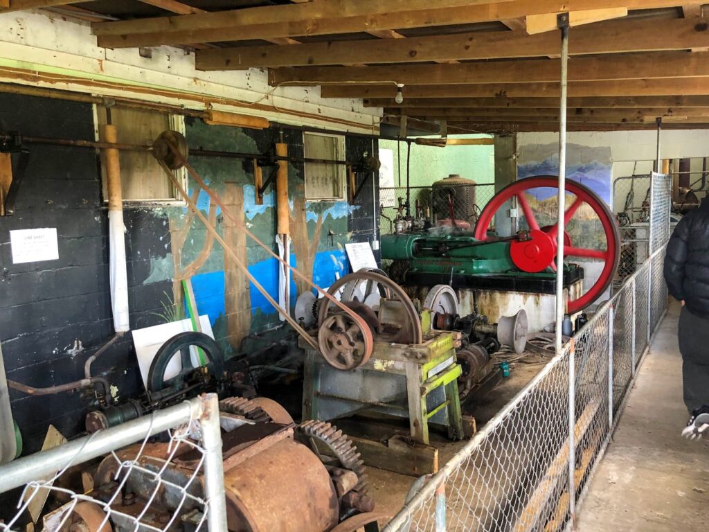 Historic line shaft machinery display featuring a large stationary engine and belt-driven equipment at a heritage workshop.
