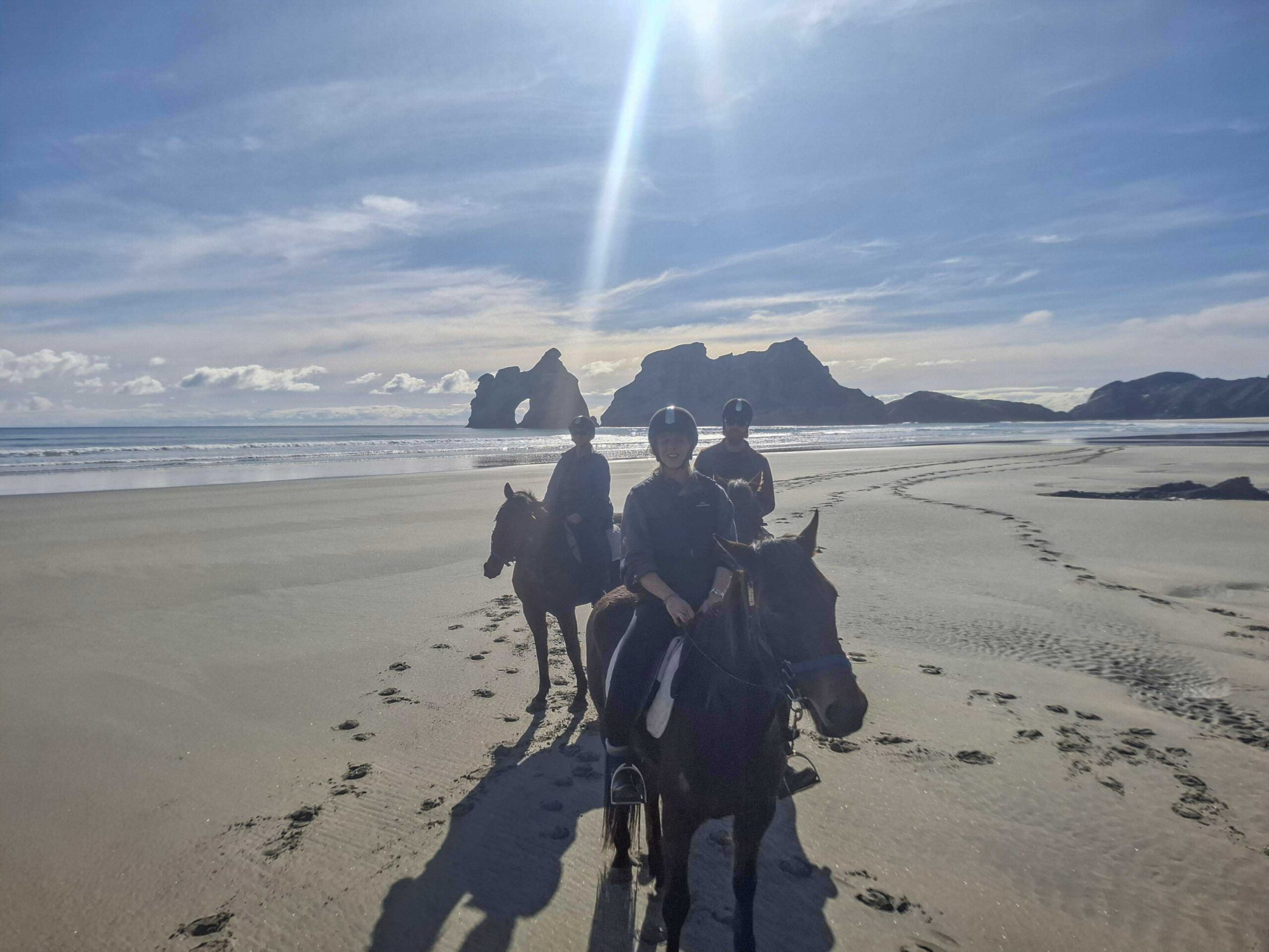 Riders on horseback enjoying a scenic beach trek at Wharariki Beach, Golden Bay, New Zealand, with the iconic Archway Islands in the background under a sunny blue sky.