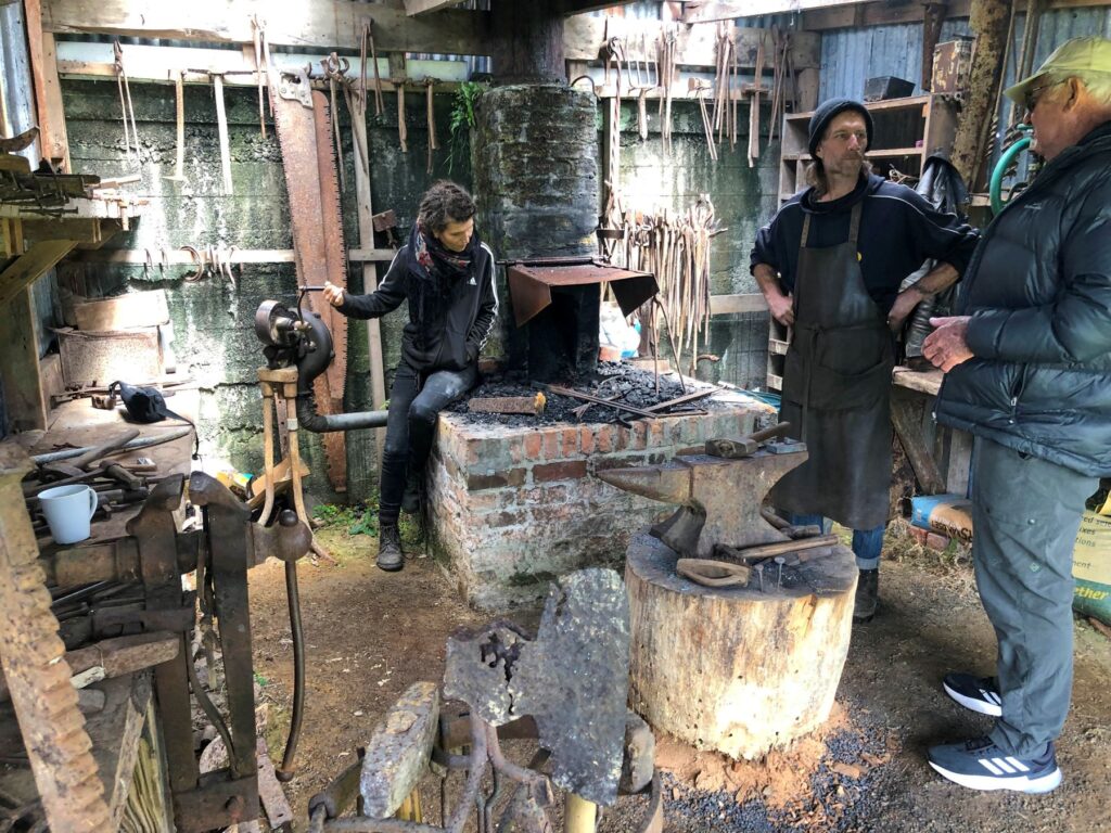 Rustic blacksmith workshop with three people gathered around a brick coal forge and an anvil on a wooden stump, surrounded by hanging hand tools.