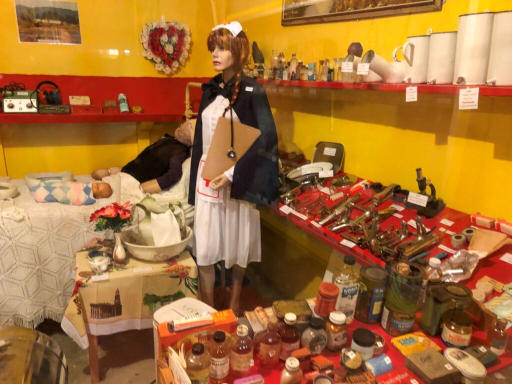 A vintage medical display featuring a mannequin dressed as a nurse, a sleeping mannequin baby, and various old medical instruments and bottles in a yellow and red-themed room.