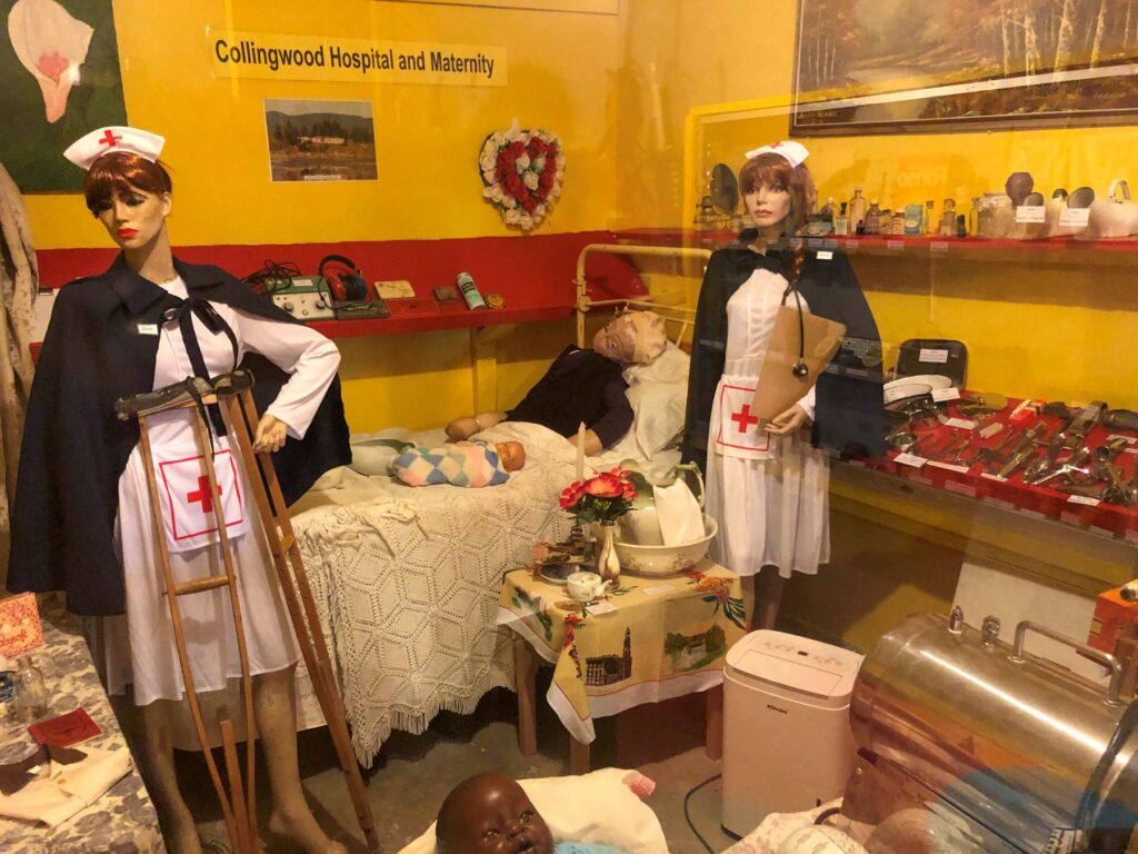 A vintage medical display at Collingwood Hospital and Maternity featuring two mannequins dressed as nurses with crutches and stethoscopes, a mannequin baby in a crib, and various antique medical tools and bottles.